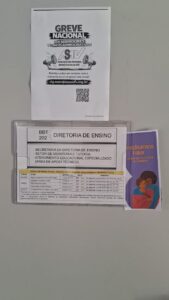 Cartazes fixados numa porta bege. O de baixo tem uma tabela com o nome da Diretoria de Ensino; o de cima traz o texto Greve Nacional dos TAEs, ASAV. À direita do cartaz, há um folder de fundo roxo com três mulheres desenhadas. 