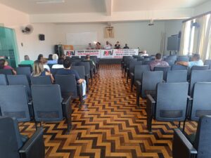 Auditório com piso de tacos de madeira e cadeiras pretas; um pequeno número de pessoas está sentado nelas. Ao fundo, uma longa mesa está coberta com faixas da Asav e, atrás delas, há 5 pessoas sentadas. Ao centro, um homem negro e robusto fala ao microfone. Na parede atrás deles há um grande crucifixo envolto por uma pequena cortina. 