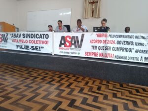 Longa mesa coberta por faixas com a logo da Asav. Atrás delas, estão sentados 4 homens e uma mulher. Um homem negro fala ao microfone, e a mulher digita em um laptop. 