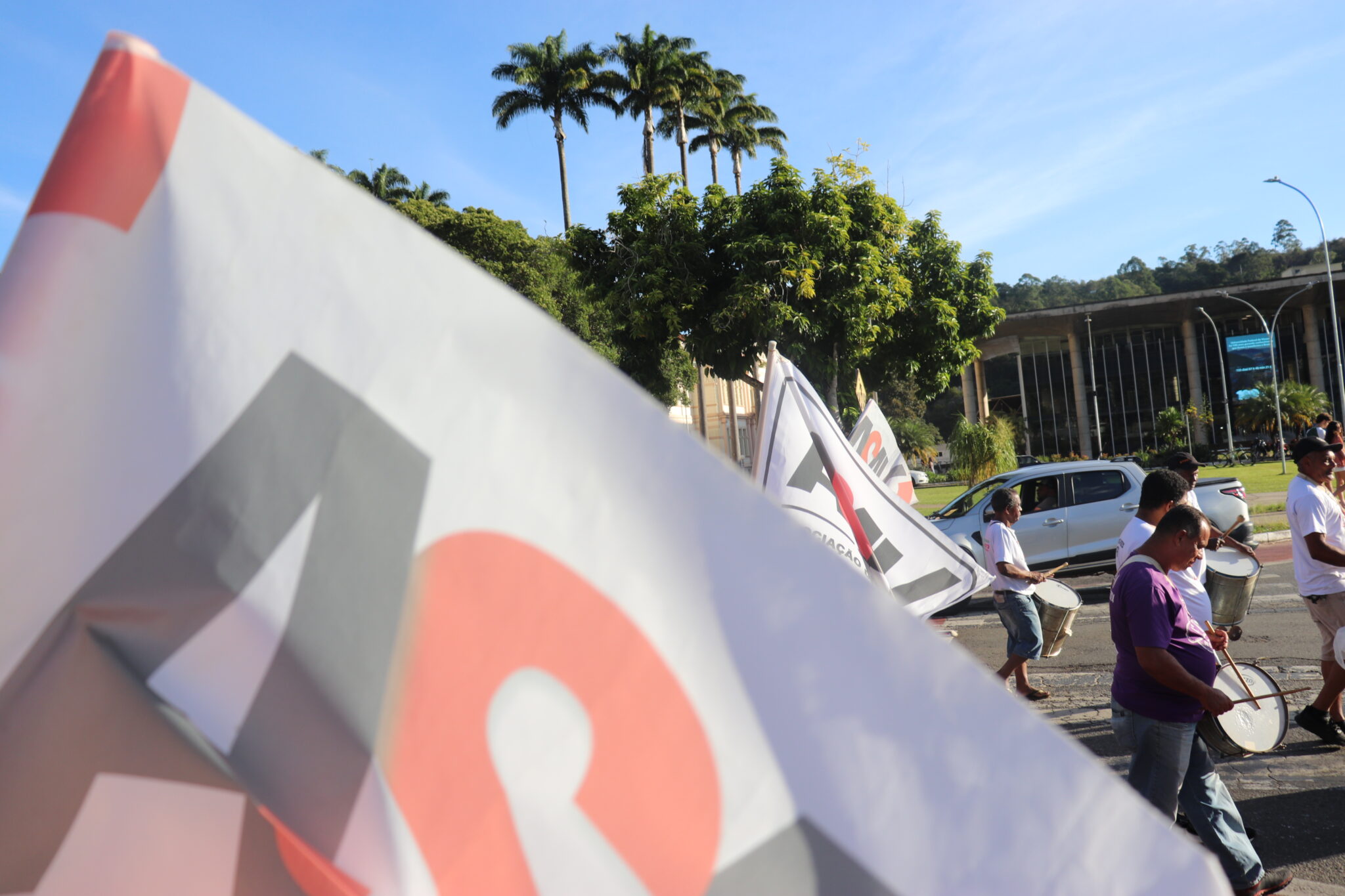 Foto em local aberto com muitas árvores e dia com céu aberto. Muitas bandeiras brancas com a logo da Asav estão sendo agitadas no alto, uma delas bem próxima de nós.