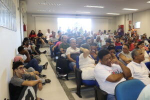 Auditório ocompletamente lotado, com pessoas de pé ao fundo. Destaque para o corredor lateral à esquerda, com jovens sentados no chão e apoiados na parede. 
