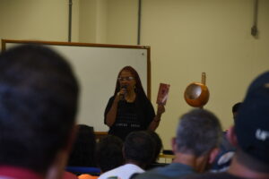 Mulher negra de cabelos longos trançados e vermelhos, óculos e camiseta preta, fala ao microfone em frente à plateia. Ela segura um folder com a mão direita. 