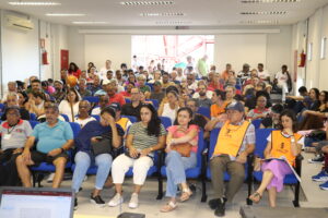 Dezenas de pessoas, de várias etnias, gêneros e idades, estão sentadas em um auditório, que tem uma grande porta nos fundos. 