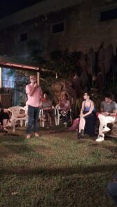 Homem de meia-idade, camisa rosa, óculos e boina colorida fala ao microfone, observado por várias pessoas ao redor. O local é uma área externa gramada, de noite. 