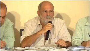 Print do vídeo em que Jaime da Silva Oliveira fala ao microfone. Ele e um homem idoso, branco, magro e calvo. 