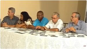Print do vídeo mostra Sebastião, Vanda, Evaristo, Frederico e Carlindo. Eles estão sentados à mesa com várias fotos espalhadas em frente a eles. 