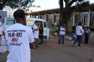 Homem branco de estatura baixa, boné e óculos fala ao microfone ao lado de uma kombi branca com a porta aberta. Ele é observado por várias pessoas que estão ao redor. 