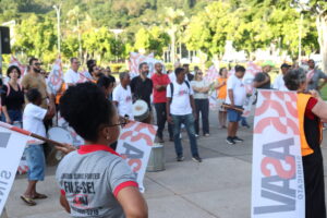 Dezenas de pessoas de pé em uma área externa cimentada, olhando para a direita, algumas com bandeiras com a logo da Asav. Perto de nós, uma mulher negra com o cabelo preso no alto da cabeça, óculos e camiseta cinza com apalavra "Filie-se" e a logo da Asav logo abaixo. 