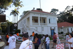 Dezenas de pessoas com bandeiras e tambores estão de pé em frente a uma casa branca de dois andares com uma grande varanda no piso superior. 