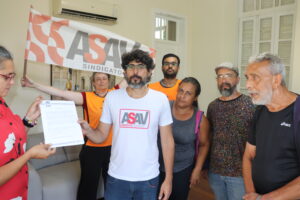 Homens e mulheres posam para a foto de maneira desorganizada. Ao centro, um homem branco, alto e magro de camiseta branca com a logo da Asav segura um documento, junto com uma mulher alta, branca, cabelos levemente grisalhos e cacheados. Atrás deles, uma mulher de meia-idade branca e colete laranja segura uma bandeira com a flogo da Asav. 