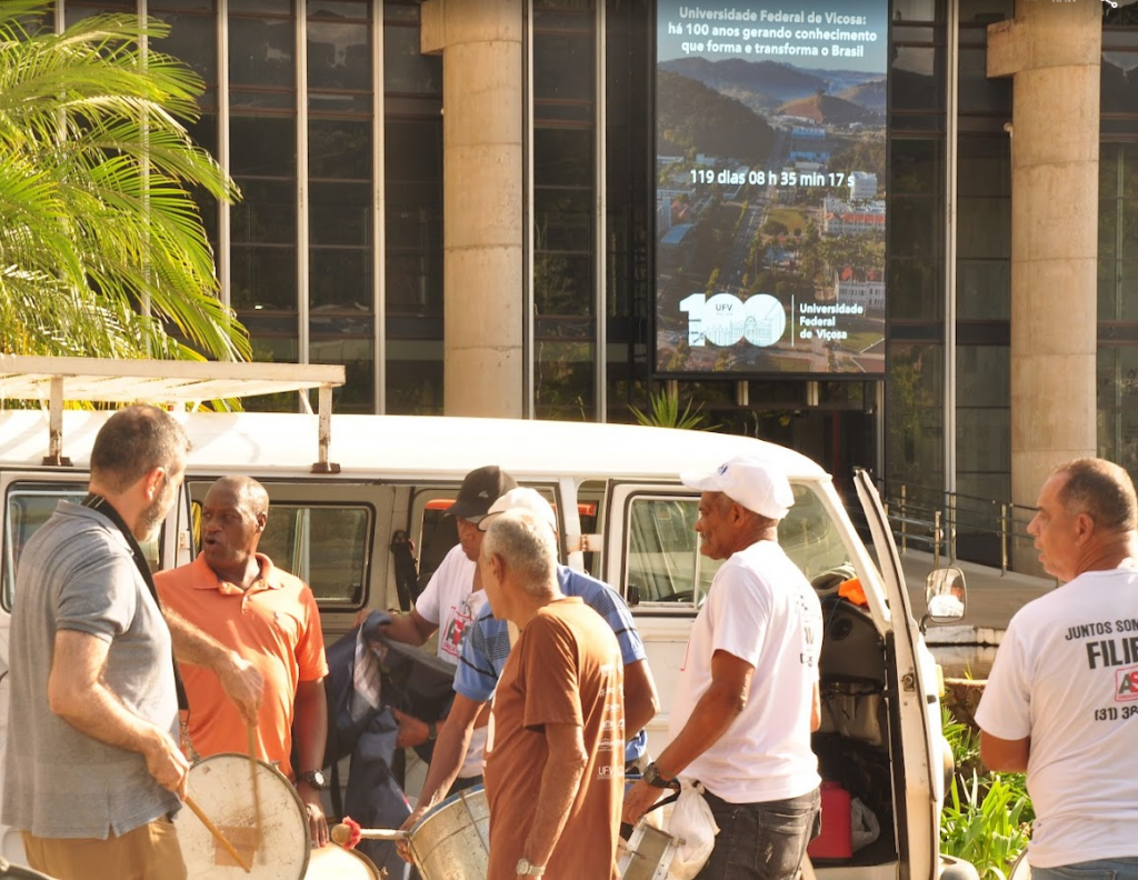 Várias pessoas retirando instrumentos de percussão de uma kombi branca, parada em frente a um prédio com fachada de vidro e grandes colunas de cimento. Nessa fachada, há um grande telão onde se lê: "Universidade Federal de Viçosa: há 100 anos gerando conhecimento que forma e transforma o Brasil"; Abaixo do texto, há um cronômetro que informa: 119 dias, 8 horas e 35 minutos.