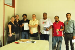 Uma mulher e dois hoomens brancos, um homem negro e um homem pardo, nessa ordem, lado a lado para a foto. Dois deles, seguram juntos uma folha de papel em frente ao corpo e sorriem. 