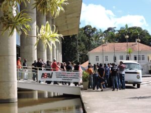 Passarela do Centro de Vivência vista de lado, passando sobre um espelho d'água. Sobre ela há dezenas de pessoas, todas olhando para uma kombi branca estacionada em frente a elas. Perto da kombi há um grande grupo de pessoas. Ao fundo, uma casa branca de dois andares, com janelas estreitas e altas. 