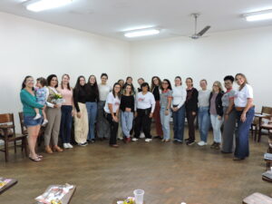Várias mulheres de diferentes idades e etnias, todas de pé, lado a lado, olhando para nós e sorrindo. A primeira da esquerda carrega uma menininha no colo, com vestidinho azul claro. Elas estão em uma grande sala de paredes brancas e piso de tacos de madeira. 