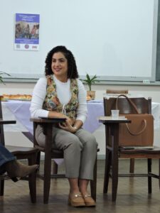 Raíssa Mourão: mulher jovem, pele clara, cabelos pretos cacheados na altura dos ombros. Ela usa blusa branca de manga longa por baixo de um colete colorido, e está sentada olhando para a direita. Ela sorri. 