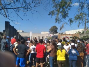 Centenas de pessoas aglomeradas em frente à entrada de um prédio baixo branco, com um letreiro "Hospital Universitário". Elas estão de costas.