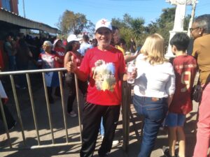 Manifestante de camisa vermelha e boné branco, de pé em frente a um gradil e virado para nós. Ele sorri e mostra os polegares das mãos. Dezenas de pessoas estão ao redor.
