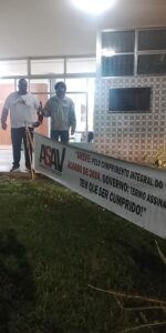 Foto vertical tirada à noite, em frente ao departamento de Economia Rural. Dois homens altos de meia-idade, , um negro e robusto e o outro branco e magro, estão parados de pé lado a lado acenando na nossa direção, próximos a uma faixa de lona com a logo da Asav e os dizeres: "greve pelo cumprimento integral do acordo de greve 2024. Governo: termo assinado tem que ser cumprido!".