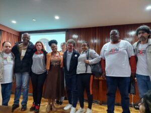 Ao centro da foto, Reginaldo Lopes está de pé, rodeado por 4 mulheres e 4 homens. Estão sobre um palco de madeira.