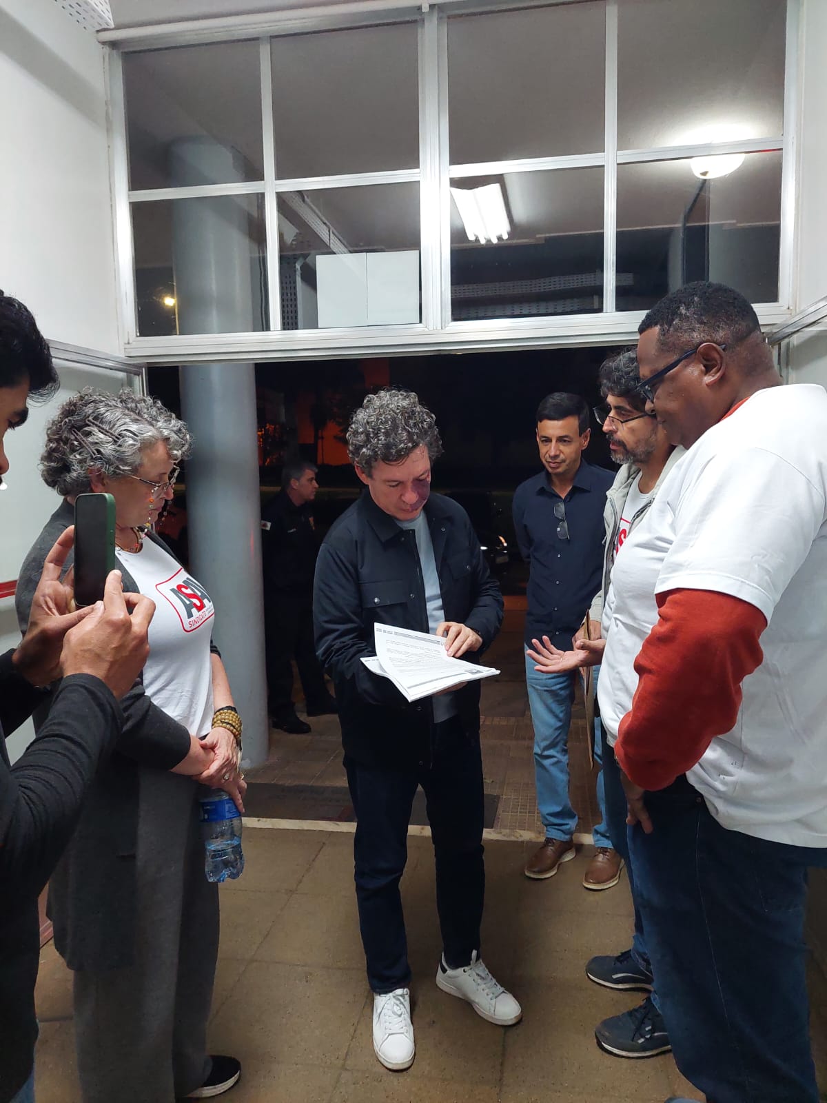 Reginaldo Lopes está ao centro da foto, rodeado por várias pessoas. Ele manuseia um documento de várias páginas.