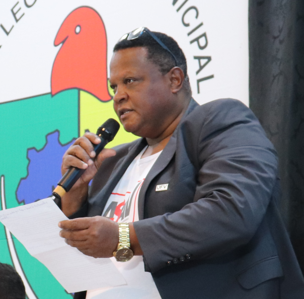 Evaristo falando ao microfone, segurando uma folha com a mão esquerda e um microfone com a direita. Ele olha para a esquerda, usa camisa branca com a logo da Asav e, por cima, paletó de terno azul escuro.