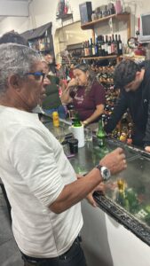 Zé Ferrujo, de pé e de frente para um freezer horizontal com tampo de vidro, com algumas garrafas de cerveja em cima. Outras pessoas estão ao redor olhando para ele e sorrindo, enquanto ele parece falar alguma coisa.