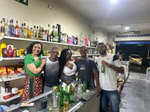 No interior de uma venda de cachaças, com prateleiras repletas de garrafas e próximo a um freezer horizontal, está Zé Ferrujo, ao lado de duas mulheres e dois homens.