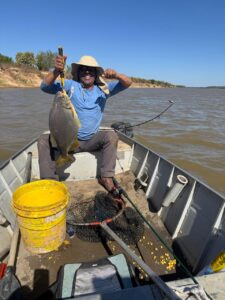 Zé Ferrujo, num barco pequeno em um rio de águas barrentas. Ele segura um peixe cinza escuro com a mão esquerda e segura o chapéu de palha em sua cabeça com a outra mão, sorridente.