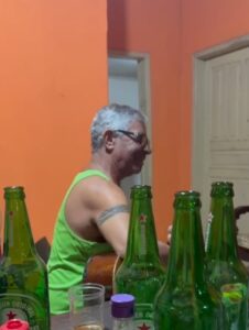 José Bhering sendado ao lado de uma mesa repleta de garras de cerveja vazias. Ele usa regata verde claro e segura um violão em frente ao corpo. Para acionar o vídeo, clique nesta imagem. 