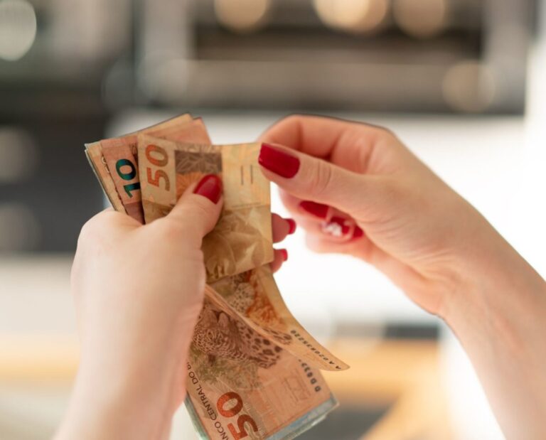 Mãos de uma mulher branca, com unhas pintadas de vermelho, contando notas de dinheiro. Aparecem mais claramente uma nota de 50 reais e outra de 10 reais logo atrás.