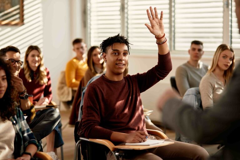 Sala de aula com estudantes adultos de diferentes gêneros e etnias. Ao centro, um homem negro jovem com blusa vermelha está com a mão erguida.